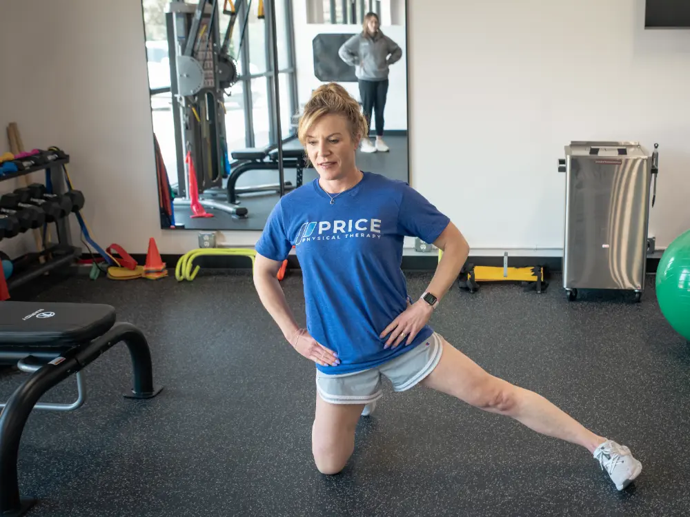 A woman in a blue "Price Physical Therapy" shirt and shorts kneels on one knee, stretching her other leg to the side in a gym to improve her balance, with exercise equipment and another person visible in the background.