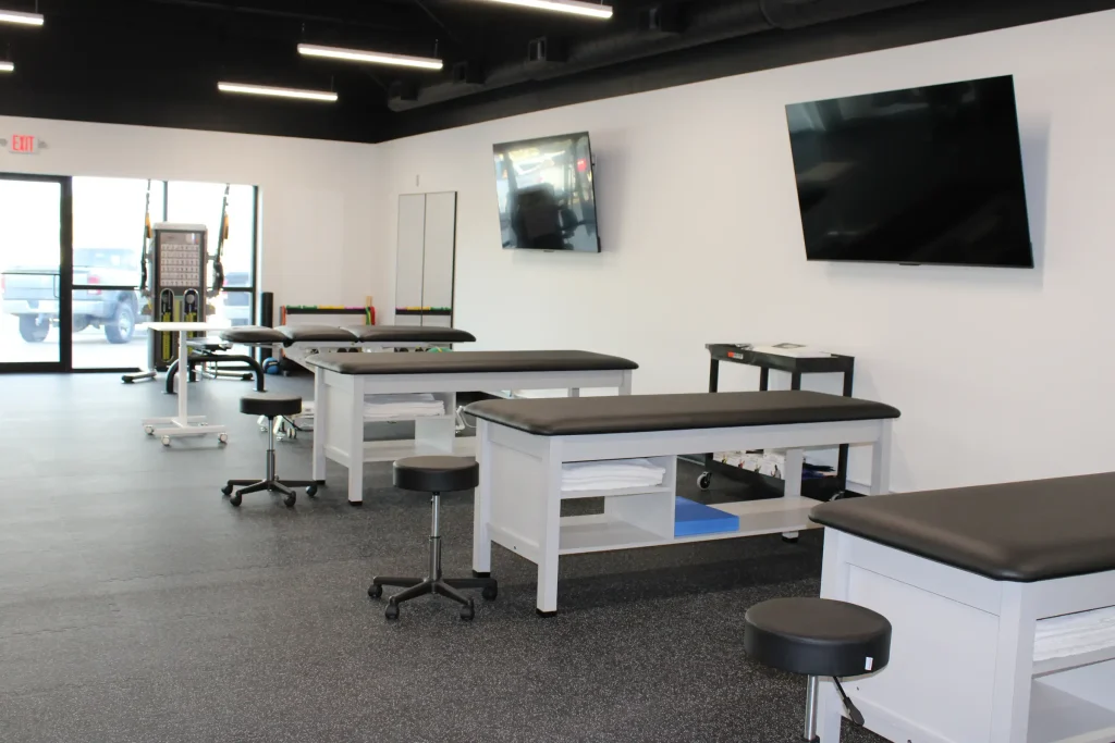 Interior of a modern physical therapy clinic featuring several therapy beds, stools, and various rehabilitation equipment, with a clean black and white color scheme.