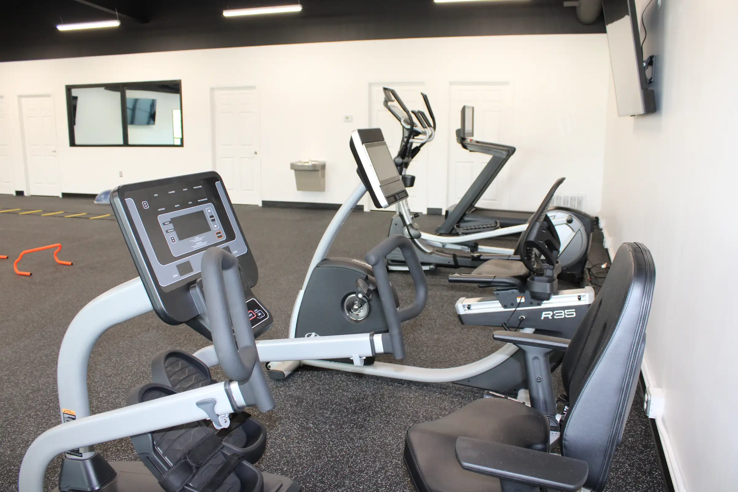 An indoor gym with several elliptical machines and recumbent bikes equipped with digital monitors, set on a grey carpeted floor against white walls with doors in the background.
